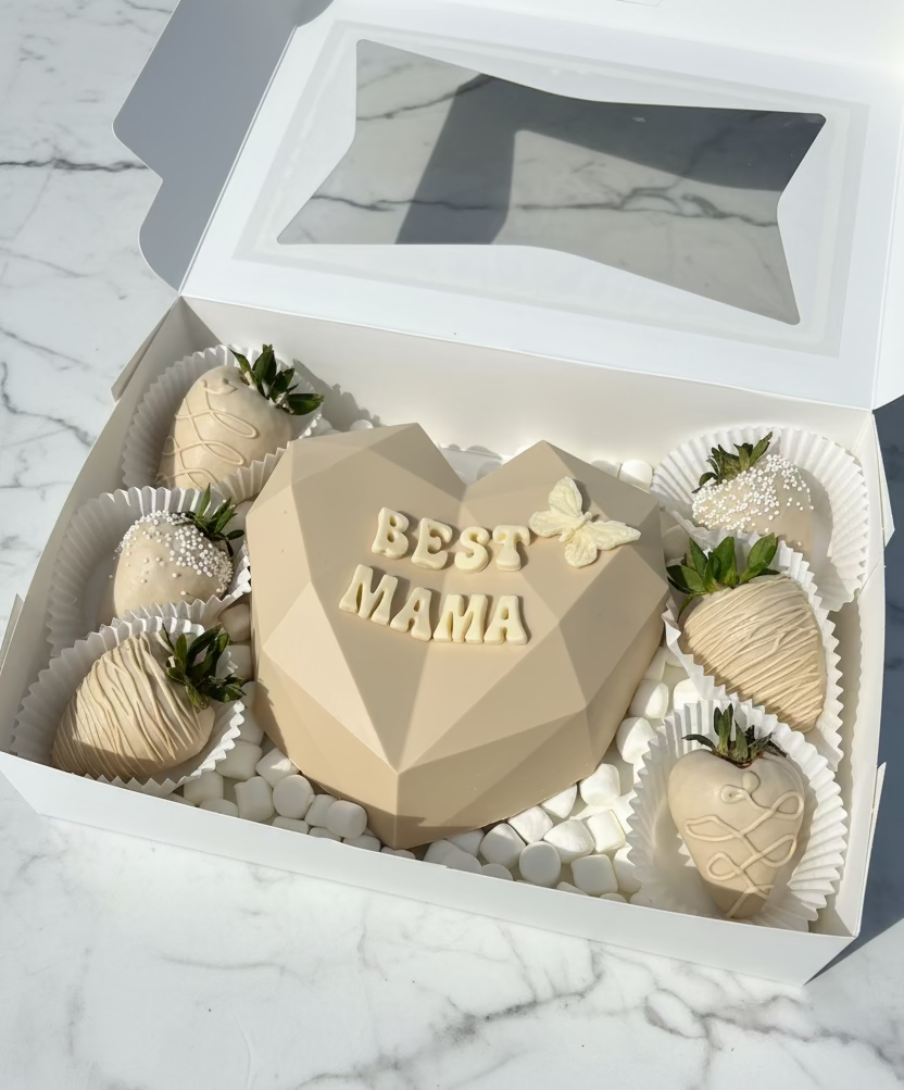 Heart-shaped cake with 'BEST MAMA' text surrounded by strawberries on a white tray outdoors.
